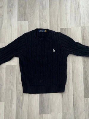 Stickad tjocktröja Polo Ralph Lauren - Kabelstickad svart tjock tröja från Polo Ralph Lauren med rund halsringning och klassisk vit broderad logga på bröstet. Tröjan har ribbade muddar vid ärmslut och nederkant. Perfekt för en stilren och tidlös look.