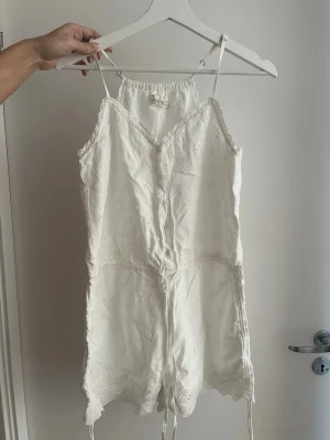 Vit spetsjumpsuit med broderi - Söt vit jumpsuit med tunna axelband, knappar framtill och broderade blommor. Har spetsdetaljer längs kanterna och dragsko i midjan. Kort modell i lätt bomullstyg, perfekt för sommardagar. Ger en romantisk och avslappnad vibe.