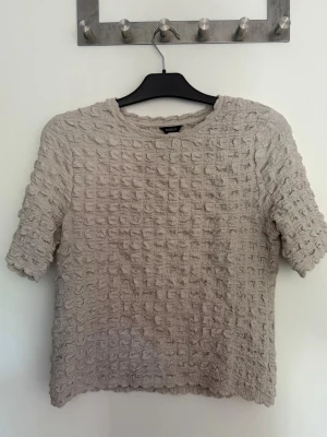 Beige topp från Lindex med struktur - Säljer en beige topp från Lindex i storlek M. Toppen har en unik strukturerad yta och rund halsringning. Materialet känns mjukt och luftigt, perfekt för en chill och stilren look. Halvlånga ärmar och enkel design gör den lätt att matcha med allt.