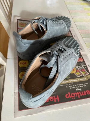 Gråa Louboutin sneakers med nitar - Säljer ett par gråa sneakers från Christian Louboutin i mocka med ikoniska röda sulor och coola nitar framtill. Skorna har matchande grå skosnören och en snygg, låg profil. Perfekta för dig som vill sticka ut med en edgy och lyxig stil. Bara använt dessa ett fåtal gånger. Hör gärna av dig vid frågor!