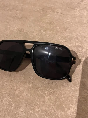 Svarta Tom Ford solglasögon - Snygga svarta solglasögon från Tom Ford med klassisk pilotform och bred båge. Glasen är mörka och bågarna har en cool detalj med öppning vid näsryggen. Perfekta för dig som vill ha en stilren och trendig look.
