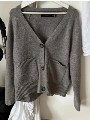Kofta från Vero Moda - Mysig kofta från Vero Moda med stora framfickor och tre knappar framtill. Koftan har ribbade muddar vid ärmslut och nederkant samt en avslappnad passform. Perfekt att slänga på sig över en t-shirt eller topp.