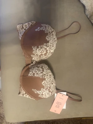 Brun push up-bh med spets från H&M - Snygg push up-bh från H&M i brun färg med vita spetsdetaljer över kuporna. Bh:n har justerbara axelband och klassisk knäppning bak. Materialet är en mix av mjuk mesh och spets som ger en romantisk look.