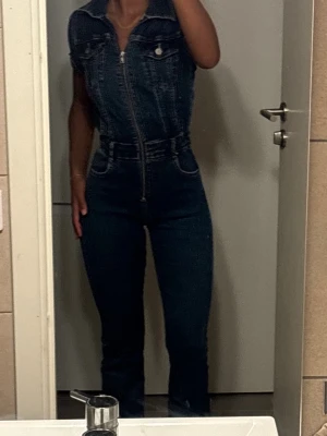 Mörkblå jeansjumpsuit med dragkedja - Snygg mörkblå jeansjumpsuit med korta ärmar, markerad midja och dragkedja framtill. Har bröstfickor med knappar och klassiska jeanssömmar. Perfekt för dig som vill ha en cool och trendig look i denim.
