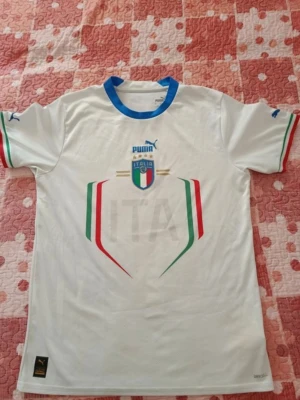 Italien borta tröja 2022 - Snygg vit fotbollströja från Puma med Italiens landslagsemblem på bröstet och blå, röda och gröna detaljer. Blå krage och Puma-logga på ärmarna. Tillverkad i lätt och ventilerande material, perfekt för träning eller match.