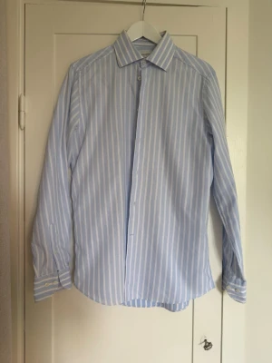 Blåvit randig skjorta med knappar - Klassisk blå och vit randig skjorta med lång ärm och knäppning framtill. Skjortan har en stilren krage och manschetter med knappar. Perfekt för en clean och fräsch look. Tillverkad i bomull för en skön känsla.
