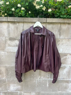Vinröd oversized jacka i läderimitation från Lindex - Snygg vinröd oversized skinnjacka från Lindex med avslappnad passform, bred krage och två framfickor. Jackan har knappar framtill och vid ärmsluten samt fina rynkdetaljer på axlar och rygg. Perfekt för dig som vill ha en unik och trendig look.