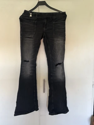 Svarta bootcut jeans med slitningar - Säljer ett par svarta bootcut jeans med slitningar på låren och knäna. Jeansen har en låg midja, fickor både fram och bak samt en lätt tvättad look. Perfekta för dig som gillar en avslappnad och trendig stil.