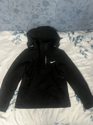 Svart Nike vindjacka med huva - Snygg svart vindjacka från Nike med huva och dragkedja framtill. Jackan har en minimalistisk design med Nike-logga på bröstet och justerbar huva. Perfekt för blåsiga dagar och enkel att matcha med olika outfits.