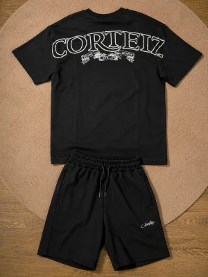 Äkta Svart set från Corteiz med tryck - Säljer ett svart set från Corteiz med t-shirt och shorts. T-shirten har stor vit logga och tryck på ryggen, medan shortsen har en liten logga på benet. Båda plaggen är i mjukt material och har en avslappnad passform. Perfekt för en chill och trendig look.
