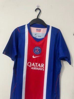 PSG matchtröja blå/röd Nike - Snygg PSG matchtröja i blått, rött och vitt från Nike. Tröjan har klubbmärke och Qatar Airways-logga på bröstet samt vita och röda ränder framtill. Tillverkad i lätt och ventilerande material, perfekt för fotboll eller träning.