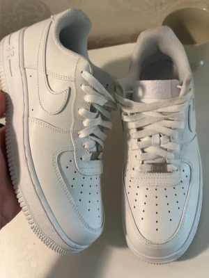Nike Air Force 1 vita sneakers - Klassiska vita Nike Air Force 1 sneakers i lågt utförande. Skorna har ovandel i slätt läder, perforeringar på tån för ventilation och en tydlig Nike Swoosh på sidan. Tjock vit sula och vita skosnören ger en clean och tidlös look.