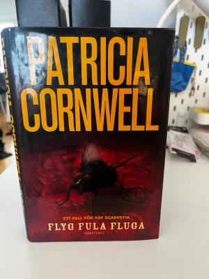 Flyg fula fluga av Patricia Cornwell - Spännande kriminalroman av Patricia Cornwell, inbunden med svart och röd omslagsdesign och stor gul text. Boken handlar om rättsläkaren Kay Scarpetta och är den tolfte delen i serien. Perfekt för dig som gillar deckare med rättsmedicinska inslag.