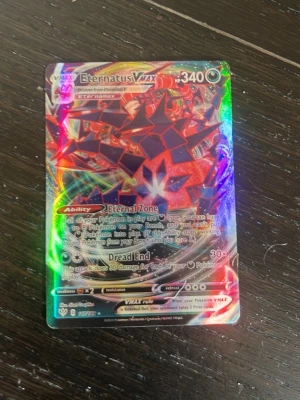 Pokémonkort Eternatus VMAX holo - Säljer ett Pokémonkort av typen Eternatus VMAX med holografisk yta. Kortet har färger i svart, rött och regnbågsskimrande detaljer. Det är tillverkat i laminerad kartong och har motiv från Pokémon Sword & Shield-serien. Inte helt säker på om den är äkta eller inte. Går att sänka priset.