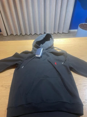 Svart hoodie från Polo Ralph Lauren - Svart hoodie från Polo Ralph Lauren med klassisk huva och dragsnören. Har en röd broderad logga på bröstet och en stor magficka framtill. Tillverkad i mjukt bomullsmaterial som är skönt mot huden. Perfekt för en avslappnad och stilren look. Ej använd 
