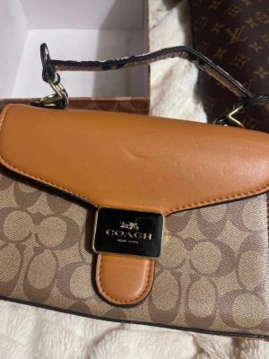 Brun handväska från Coach - Snygg handväska från Coach i klassiskt monogrammönster med ljusbruna och mörkbruna toner. Väskan har ett lock i slätt läder, guldfärgade metalldetaljer och ett kort handtag. Perfekt för dig som gillar stilrena accessoarer med lyxig känsla.