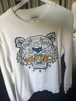 Vit Kenzo sweatshirt med tigertryck - Säljer en vit sweatshirt från Kenzo med det ikoniska tigerhuvudet broderat framtill och texten 'KENZO PARIS' i blått och guld. Tröjan har rund halsringning och ribbade muddar. Perfekt statement-plagg för dig som gillar streetwear och snygga detaljer.