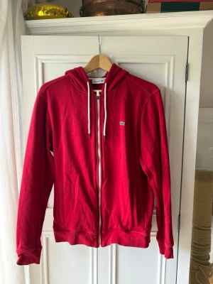 Röd hoodie med dragkedja från Lacoste - Snygg röd hoodie från Lacoste med dragkedja framtill och vita dragsnören i huvan. Klassisk krokodil-logga på bröstet och två fickor fram. Tillverkad i mjukt bomullsmaterial som känns skönt mot huden. 