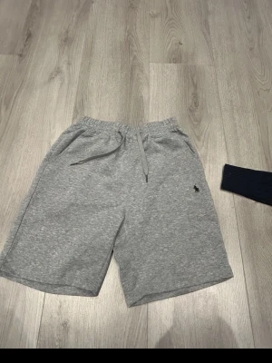 Grå shorts från Polo Ralph Lauren - Snygga grå shorts från Polo Ralph Lauren med klassisk logga broderad på benet. De har elastisk midja med snörning och är tillverkade i mjuk bomull, perfekta för en avslappnad stil.