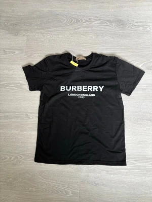 Svart Burberry t-shirt med tryck - Snygg svart t-shirt från Burberry med klassiskt vitt tryck på bröstet där det står 'BURBERRY LONDON ENGLAND PARIS'. T-shirten har rund hals och korta ärmar, perfekt för en clean och stilren look.