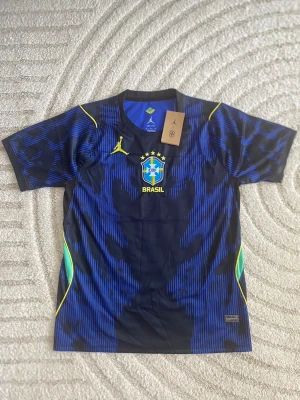 Blå Brasilien matchtröja från Jordan - Snygg blå matchtröja från Jordan i samarbete med Brasilien fotbollslandslag. Tröjan har ett mönster i olika blå nyanser, gula detaljer och Brasil-logga på bröstet. Tillverkad i lätt och ventilerande material, perfekt för träning eller match.