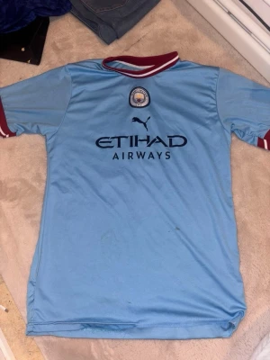 Manchester City matchtröja Haaland - Ljusblå Manchester City matchtröja från Puma med HAALAND och nummer 9 på ryggen. Tröjan har klubbmärke och Etihad Airways-logga på bröstet samt vinröda detaljer vid krage och ärmslut. Perfekt för fotbollsfans som vill stötta sitt lag.
