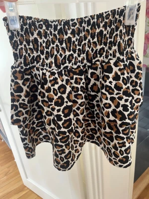 Kort leopardmönstrad kjol - Säljer en kort kjol med leopardmönster i svart, brun och beige. Kjolen har en bred, smockad midja som ger en bekväm och stretchig passform. Materialet känns lätt och luftigt, perfekt för varma dagar. Passar dig som vill sticka ut med ett djuriskt print!