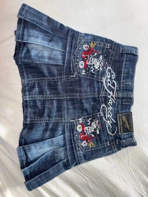 Mörkblå jeanskjol från Ed Hardy - Säljer en mörkblå jeanskjol från Ed Hardy med plisserad nederdel, nitar och broderade detaljer på bakfickorna. Kjolen har coola broderier med motorcyklar och logga, samt små pärlor framtill. Perfekt för dig som gillar Y2K och statement-plagg. 📀🖤