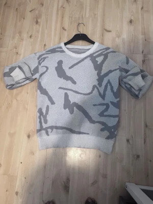 Gråmelerad stickad tröja - Säljer en gråmelerad stickad tröja med abstrakt mönster i olika grå nyanser. Tröjan har rund halsringning, ribbade muddar och avslappnad passform. Perfekt för dig som gillar streetwear och unika prints.
