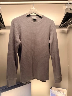 H&M - En stilren grå långärmad tröja med rund hals och waffle-mönstrad struktur. Perfekt för lager-på-lager och enkel att matcha med jeans eller chinos. Tröjan har ribbade muddar vid ärmslut och nederkant för en snygg passform.