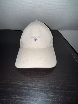 Beige keps från GANT nästan oanvänd  - Stilren beige keps från GANT med böjd skärm och broderad logotyp framtill. Klassisk sexpanelsdesign och justerbar rem baktill. Perfekt accessoar för en avslappnad och sportig look.