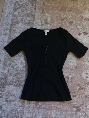 Svart ribbad topp med knappar - Snygg svart ribbad topp med korta ärmar och rundad halsringning. Toppen har en knappdetalj framtill med fem knappar som ger en stilren look. Perfekt att matcha med jeans eller kjol för en enkel men trendig outfit.