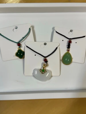 Set med tre halsband med jadeinspirerade hängen - Säljer ett set med tre halsband, alla med olika jadeinspirerade hängen. Ett halsband har ett grönt klöverformat hänge, ett har ett vitt halvmåneformat hänge och ett har ett droppformat grönt hänge. Alla hängen har guldfärgade detaljer och röda pärlor. Perfekt för dig som gillar unika smycken.