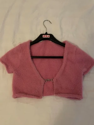 Rosa fluffig kortärmad kofta - Supersöt rosa fluffig kofta med korta ärmar och croppad passform. Koftan har en öppen front med en liten metallhake som stängning. Perfekt för att lägga till en gullig touch till din outfit.