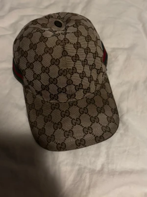 Gucci beige keps med GG-mönster - Säljer en beige keps från Gucci med det klassiska GG-monogrammönstret över hela kepsen. På sidan finns en grön och röd rand som ger extra lyxig känsla. Kepsen har böjd skärm och är tillverkad i canvasmaterial. Perfekt för dig som vill ha en stilren och trendig accessoar. Bytt stil så hade gärna blivit av med den snabbt, kan gå ner i pris!!!!