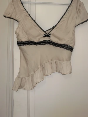 Beige topp med spets och volang - Söt beige omlottopp med svart spetsdetalj vid urringning och midja samt liten rosett framtill. Toppen har korta ärmar, v-ringning och asymmetrisk volangkant nedtill. Perfekt för dig som gillar romantiska detaljer och unik design.