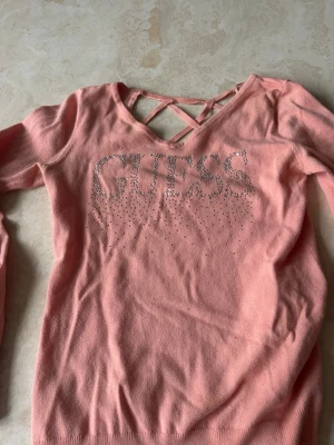 Rosa tröja med strass från Guess - Supersnygg rosa tröja från Guess med strassdetaljer som bildar loggan på framsidan. Tröjan har långa ärmar, v-ringning och coola korsade band i ryggen för extra stilpoäng. Materialet känns mjukt och stretchigt, perfekt för en trendig look.