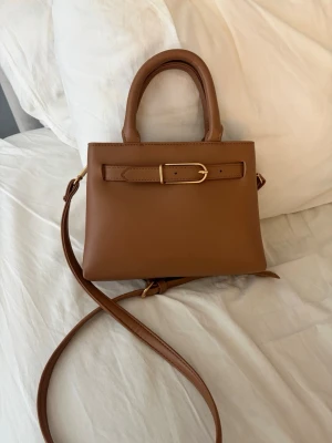 Brun handväska med guldspänne - Snygg brun handväska från Primark slätt skinn med guldfärgat spänne framtill. Väskan har både handtag och en lång axelrem för olika stilar. Perfekt storlek för det viktigaste och stängs med magnetknapp. HELT NY ALDRIG ANVÄND!
