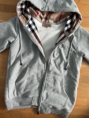 Grå hoodie från Burberry med dragkedja - Snygg grå hoodie från Burberry med dragkedja och klassiskt rutigt foder i huvan. Hoodien har två fickor framtill, ribbade muddar och justerbar dragsko i huvan. Tillverkad i mjukt bomullsmaterial för en skön och avslappnad look.
