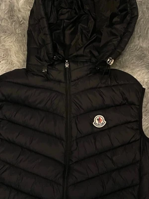 Svart dunväst från Moncler med huva - Snygg svart dunväst från Moncler med huva och dragkedja framtill. Västen har ett diskret Moncler-märke på bröstet och horisontella sömmar som ger en cool look. Perfekt att slänga över en hoodie för extra värme. Kan sänka vid snabbnaffär 