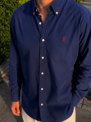 Marinblå skjorta från Ralph Lauren - Klassisk marinblå skjorta från Ralph Lauren med röd broderad logga på bröstet. Skjortan har långa ärmar, knappar framtill och button-down krage. Tillverkad i bomull för en skön och tidlös look.