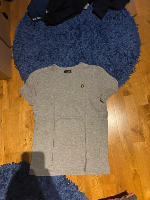 Grå t-shirt från Lyle & Scott - Snygg grå t-shirt från Lyle & Scott med klassisk gul logga på bröstet. T-shirten har rund hals och korta ärmar, tillverkad i mjuk bomull för en bekväm känsla. Perfekt basplagg till garderoben.