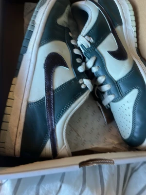 Nike Dunk Low grön/vit sneaker - Nike Dunk Low sneakers i grönt och vitt läder med klassisk svart Swoosh på sidan. Skorna har vit mellansula och grön yttersula, samt perforerad tåbox för extra ventilation. Ikonisk retrostil som passar perfekt till streetwear och casual outfits.