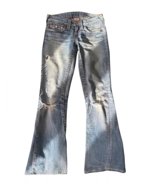 Vintage Ljusblå bootcut jeans från True Religion - Säljer ett par vintage  ljusblå bootcut jeans från True Religion med slitningar och hål på benen. Jeansen har klassiska bakfickor med lock och knappar samt orangea sömmar. Tillverkade i denim med en cool vintagekänsla. Dem är storlek w24 men de äldre modellerna är mycket mindre så passar mer w23 w22