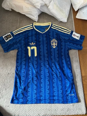 Blå Sverige fotbollströja Adidas - Säljer en blå fotbollströja från Adidas med Sveriges landslagsemblem och nummer 17 i gult. Tröjan har retroinspirerat mönster, gula detaljer och tre klassiska ränder på axlarna. Tillverkad i lätt och ventilerande material, perfekt för match eller träning.