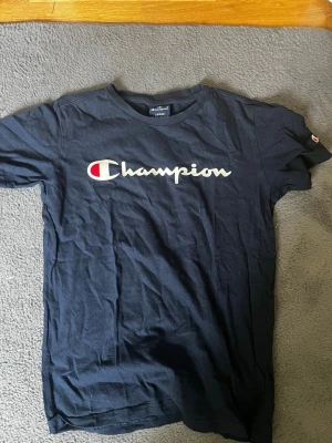 Svart Champion t-shirt med logga - Snygg svart t-shirt från Champion med klassisk logga i vitt och rött på bröstet. T-shirten har rund halsringning och korta ärmar. Perfekt till jeans eller shorts för en avslappnad streetwear-look.