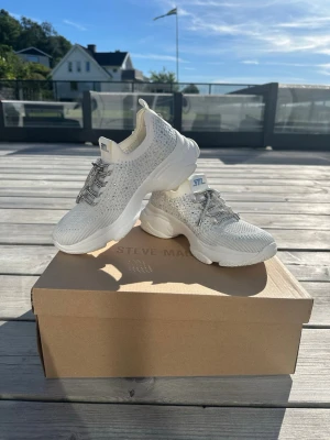 Vita sneakers med strass från STEVE MADDEN, helt nya!  - Säljer ett par vita sneakers från STEVE MADDEN med  ovandel i tyg och glittrande strassdetaljer. Skorna har tjock sula, silvriga skosnören och logga baktill. Perfekta för dig som vill sticka ut med en sportig men ändå glamorös look. Helt nya! Endast provade! Ordinarie pris: 1399:- 