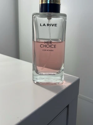 La Rive Her Choice Eau de Parfum - La Rive Her Choice Eau de Parfum för kvinnor i en elegant, rektangulär glasflaska med ljusrosa doftvätska. Flaskan har svart och guldfärgad spraykork och stilren text på framsidan. Perfekt för dig som gillar fräscha och feminina dofter.