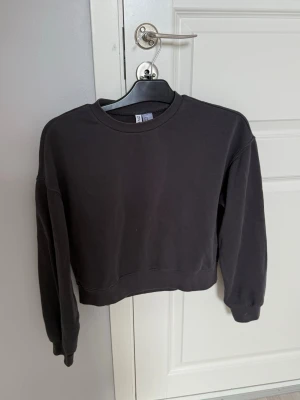Svart croppad hoodie från H&M - En enkel och stilren svart croppad hoodie från H&M. Tröjan har rund halsringning, långa ärmar och ribbade muddar vid ärmslut och nederkant. Perfekt för en avslappnad och trendig look. Tillverkad i mjukt bomullsmaterial.
