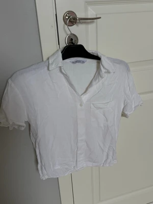 Vit croppad skjorta med kort ärm - Säljer en vit croppad skjorta med korta ärmar och klassisk krage. Skjortan har en bröstficka och knappar framtill. Perfekt för en avslappnad och trendig look.
